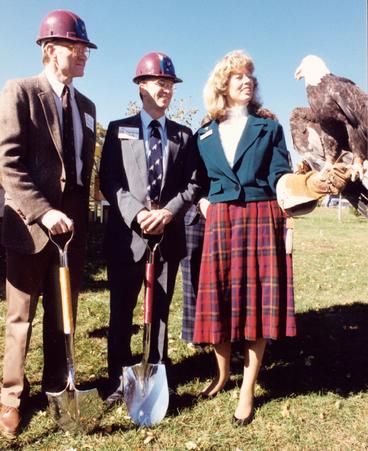 Two men with construction hats on stand next to a woman with long blonde hair with a bald eagle perched on her gloved hand